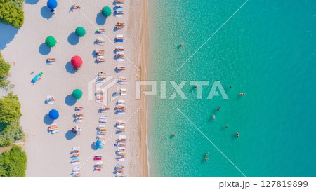 Tourists are sunbathing on a white sand beach under colorful umbrellas, while other people are swimming in the clear turquoise water, creating a vibrant summer scene 127819899