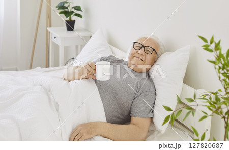 Senior elderly man lying on bed in bedroom holding a cup of coffee or tea enjoying good morning Senior elderly man lying on bed in bedroom holding a cup of coffee or tea enjoying good morning 127820687