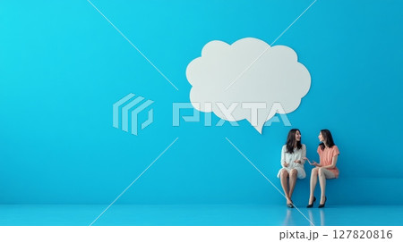 Two young women are sitting against a blue wall, having a conversation under a large blank speech bubble, symbolizing communication and brainstorming 127820816