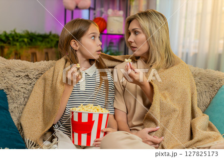 Child girl kid with mother watching cartoon together at home on sofa under warm blanket in room 127825173