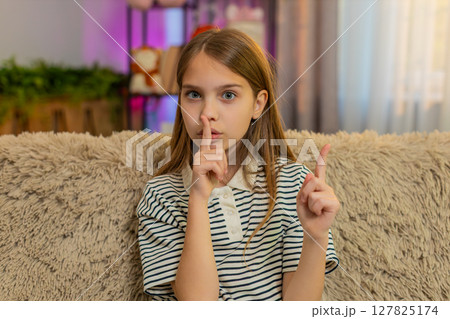 Child girl kid sitting on sofa at home showing silence gesture with finger near lips looking serious 127825174