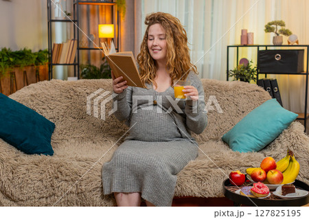 Pregnant young woman drinking orange juice while reading pregnancy book at home, calm relaxed mood 127825195