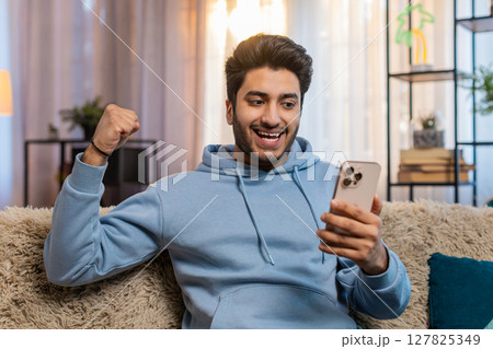 Excited Indian man holding smartphone celebrating online game win sitting on sofa in living room 127825349