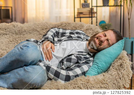 Young ill sick man lying at sofa with palm on abdomen moaning from stomach trouble or overeating 127825392