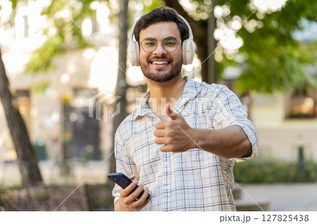 Happy Indian man in wireless headphones choosing, listening disco music on smartphone on street Happy Indian man in wireless headphones choosing, listening disco music on smartphone on street 127825438