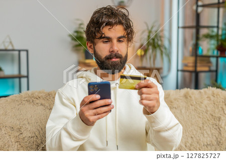 Happy young Hispanic man guy using credit bank card and cellphone while shopping online at home Happy young Hispanic man guy using credit bank card and cellphone while shopping online at home 127825727