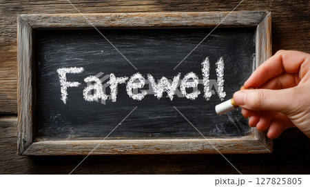 Hand holding white chalk writes Farewell on rustic chalkboard, conveying sense of goodbye 127825805