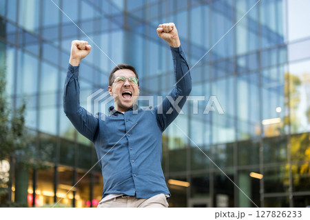 A man with glasses raises his arms in celebration in front of a modern glass building. He appears excited and happy. A man with glasses raises his arms in celebration in front of a modern glass building. He appears excited and happy. 127826233