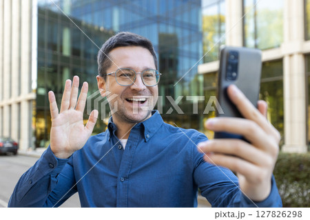 A cheerful man wearing glasses waves while holding his phone for a video call. Modern office building in the background. A cheerful man wearing glasses waves while holding his phone for a video call. Modern office building in the background. 127826298
