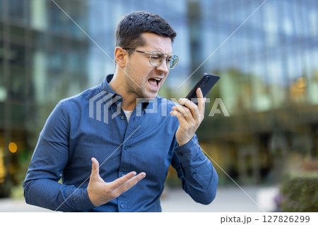 An angry businessman wearing glasses shouts on a smartphone while standing outdoors. He is visibly frustrated, gesturing with his hands. An angry businessman wearing glasses shouts on a smartphone while standing outdoors. He is visibly frustrated, gesturing with his hands. 127826299
