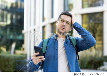 A concerned man looks at his phone while holding his head outside near a modern building. He is wearing glasses and a backpack. 127826333