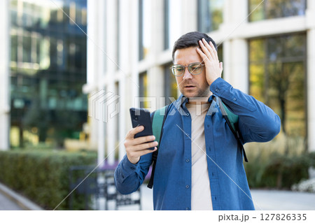 A concerned man outside is looking at his smartphone, hand on his head, possibly in response to bad news. 127826335