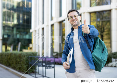 A smiling student with a thumbs-up gesture, wearing a backpack and holding a notebook, stands in front of a modern building. 127826342