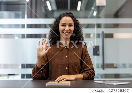 A smiling woman with curly hair waves at the camera while wearing a headset in an office setting. A smiling woman with curly hair waves at the camera while wearing a headset in an office setting. 127826367