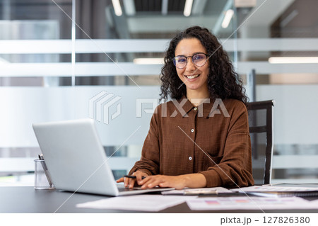 A smiling woman works on a laptop in a modern office environment, radiating professionalism and positivity. A smiling woman works on a laptop in a modern office environment, radiating professionalism and positivity. 127826380