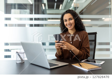 Portrait of a successful satisfied businesswoman at her workplace inside the office. The woman is working with a laptop, holding a phone in her hands, smiling and looking at the camera. Portrait of a successful satisfied businesswoman at her workplace inside the office. The woman is working with a laptop, holding a phone in her hands, smiling and looking at the camera. 127826470