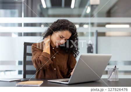 A woman with curly hair, working on a laptop, massages her neck in pain, likely from strain. 127826476