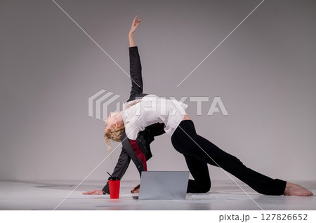 A ballerina dressed in a business suit poses for a laptop and drinks coffee. Flexible woman works at the computer A ballerina dressed in a business suit poses for a laptop and drinks coffee. Flexible woman works at the computer 127826652