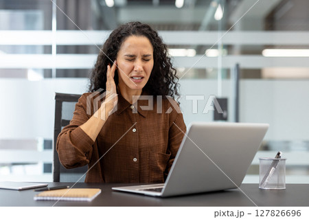 A woman with curly hair is in pain, holding her ear while working on a laptop in an office setting. 127826696