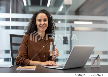 A smiling woman is holding a glass of water and working on a laptop in an office setting. A smiling woman is holding a glass of water and working on a laptop in an office setting. 127826712
