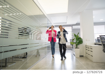 Two colleagues are walking through the office and talking on the phones. Two colleagues are walking through the office and talking on the phones. 127826728