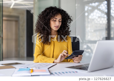 A focused woman works diligently at a desk, utilizing a calculator alongside a laptop and financial documents in a bright office. 127826827