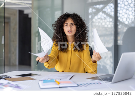 Hispanic businesswoman showing frustration holding papers at desk with laptop, notebook. Emotion conveyed through facial expression and body language. Concept of stress in professional environment. Hispanic businesswoman showing frustration holding papers at desk with laptop, notebook. Emotion conveyed through facial expression and body language. Concept of stress in professional environment. 127826842