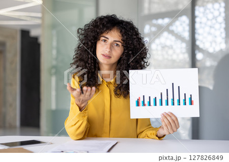 A businesswoman presents a graph, gesturing with a pen while looking at the camera in an office setting. A businesswoman presents a graph, gesturing with a pen while looking at the camera in an office setting. 127826849