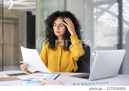 An accountant is frustrated with paperwork at her desk in the office. She is holding a pen and some documents in her hand 127826880