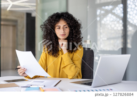 A thoughtful accountant reviews paperwork in a modern office setting, looking for discrepancies and ensuring compliance A thoughtful accountant reviews paperwork in a modern office setting, looking for discrepancies and ensuring compliance 127826884