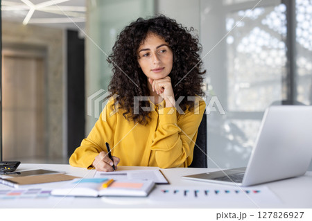 Serious and confident businesswoman working with documents and reports. Woman thinking about current financial investment tasks, preparing report, and filling tax forms while sitting inside office. 127826927