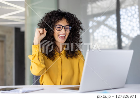 Successful joyful woman celebrating victory and successful achievement results. Businesswoman at workplace working with laptop holding hand up, gesture of triumph. 127826971
