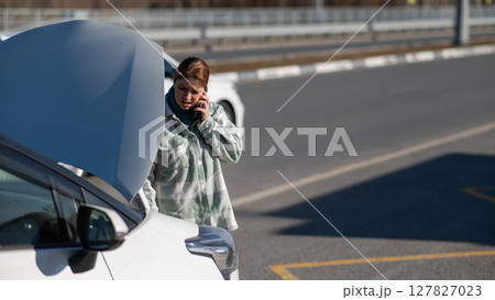 Caucasian woman cries and calls on the phone standing near the car with the hood open. Caucasian woman cries and calls on the phone standing near the car with the hood open. 127827023
