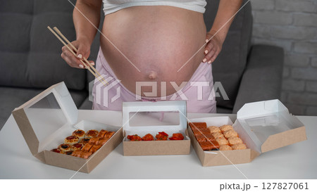 A pregnant woman holds chopsticks and stands at the table with rolls. Close-up of the belly. A pregnant woman holds chopsticks and stands at the table with rolls. Close-up of the belly. 127827061