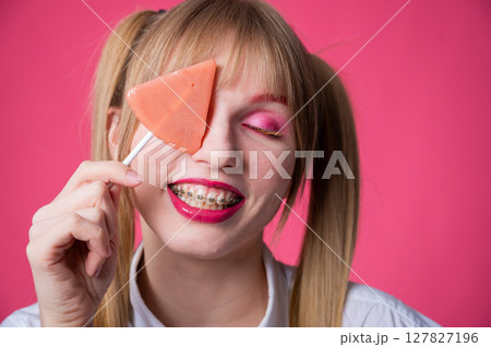 Portrait of a young woman with braces and bright makeup eating a lollipop on a pink background. Portrait of a young woman with braces and bright makeup eating a lollipop on a pink background. 127827196