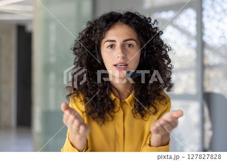 A young woman with curly hair is speaking into a headset, gesturing with her hands, and providing customer service. 127827828