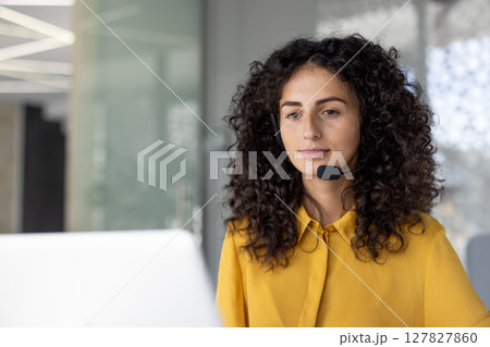 A woman with curly hair wearing a headset, looking attentively at a computer screen, likely providing customer service or support in a modern office. A woman with curly hair wearing a headset, looking attentively at a computer screen, likely providing customer service or support in a modern office. 127827860
