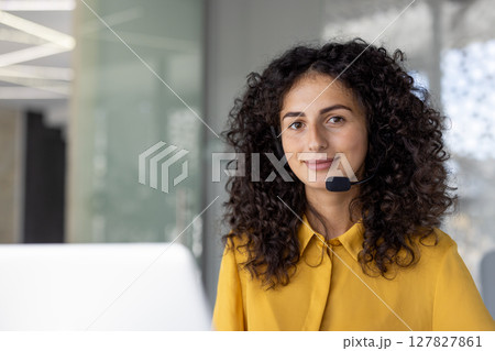 A smiling woman with curly hair wears a headset. She is likely working in a customer service or call center environment. A smiling woman with curly hair wears a headset. She is likely working in a customer service or call center environment. 127827861