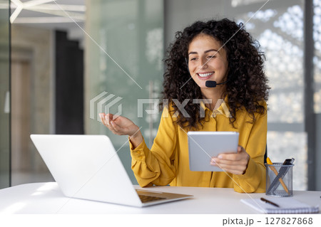 Support service worker, businesswoman with headset and tablet in hands communicates and consults clients remotely, office worker smiles satisfied uses laptop inside office. 127827868