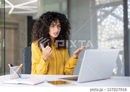 A woman with curly hair appears frustrated during a video call, gesturing with her hands while seated in front of a laptop. 127827916