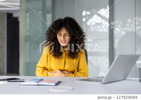 Hispanic businesswoman sits at office desk, experiencing stomach pain. She holds her abdomen, showing stress and discomfort. Nearby are laptop and notebook, indicating a work setting. 127828089