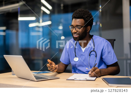 A black male doctor in scrubs and stethoscope smiles while taking notes during a telehealth appointment in his modern office. 127828143