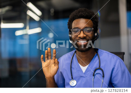 A smiling healthcare professional wearing a headset waves hello, seemingly ready to assist. The doctor sports glasses and a stethoscope. 127828154