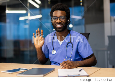 A friendly doctor in scrubs and a headset is seated at his desk and waving to greet someone during a telehealth visit. 127828158