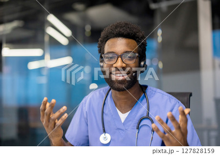 An African-American male doctor wearing glasses and a headset smiles while gesturing during a telehealth consultation. A stethoscope is around his neck. 127828159