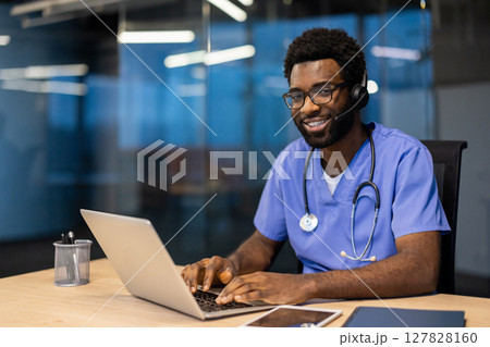 A smiling, doctor wearing glasses and a headset uses a laptop in an office setting. He has a stethoscope on and looks ready to assist patients. 127828160