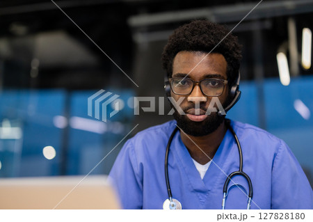 Young doctor with headset looking at camera, close-up portrait of doctor inside office. Man in blue medical coat and stethoscope working remotely, consulting clinic patients and helping with advice. Young doctor with headset looking at camera, close-up portrait of doctor inside office. Man in blue medical coat and stethoscope working remotely, consulting clinic patients and helping with advice. 127828180