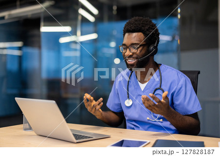 Successful experienced doctor with headset and laptop inside office at workplace working remotely. Man consulting patients, helping and supporting, video call, remote meeting conference. 127828187