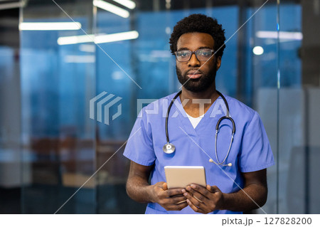 An african american doctor poses confidently with a tablet. He is wearing scrubs and eyeglasses, a stethoscope around his neck. An african american doctor poses confidently with a tablet. He is wearing scrubs and eyeglasses, a stethoscope around his neck. 127828200