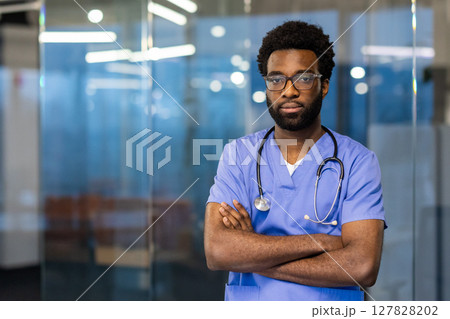 A confident medical professional in scrubs stands in a modern hospital setting with arms crossed. He wears eyeglasses and a stethoscope. A confident medical professional in scrubs stands in a modern hospital setting with arms crossed. He wears eyeglasses and a stethoscope. 127828202